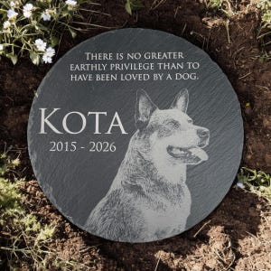 May include: A round dark gray slate memorial stone featuring an engraved portrait of a dog, the name KOTA, the years 2015-2026, and the text: There is no greater earthly privilege than to have been loved by a dog. The stone has a textured surface.