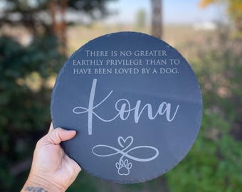 May include: A round, dark gray slate plaque featuring the engraved text "THERE IS NO GREATER EARTHLY PRIVILEGE THAN TO HAVE BEEN LOVED BY A DOG." Below this, the name "Kona" is written in script, followed by an infinity symbol with a heart and paw print.