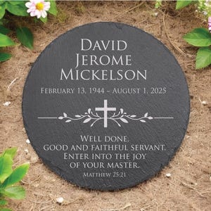 May include: A round black slate memorial stone engraved with the name David Jerome Mickelson, the dates February 13, 1944 to August 1, 2025, a cross icon with leafy branches, and the text Well done, good and faithful servant. Matthew 25:21.