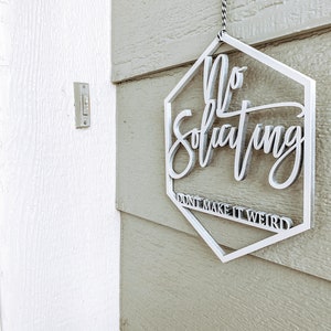 May include: White hexagon-shaped sign with the text "No Soliciting Don't Make It Weird" hanging on a light green wooden door.