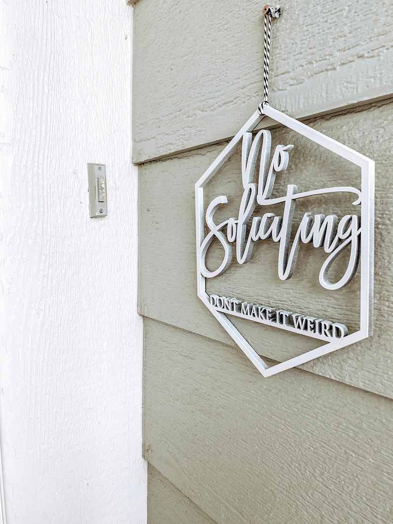 May include: White hexagon-shaped sign with the text "No Soliciting Don't Make It Weird" hanging on a light green wooden door.