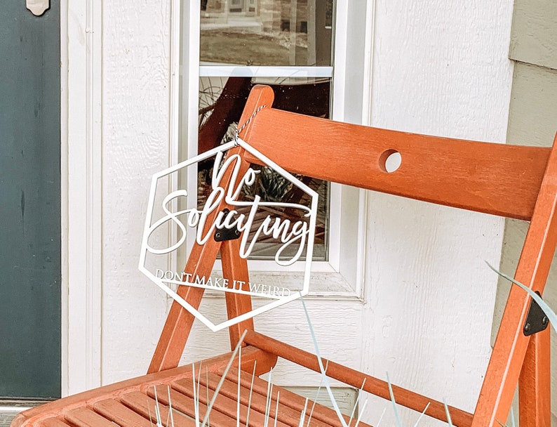 May include: White hexagon-shaped sign with the text "No Soliciting Don't Make It Weird" hanging on a wooden chair.