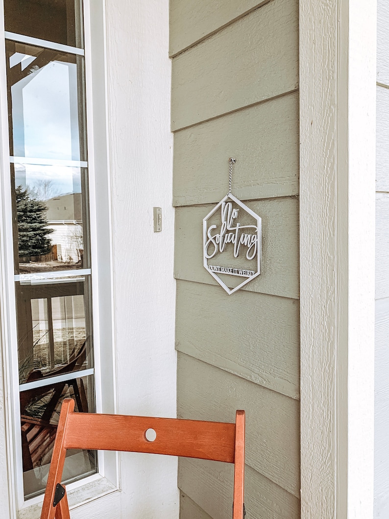May include: A silver metal sign with the text "No Soliciting" and "Just Make It Weird" hanging on a light green wooden door. A wooden chair is in the foreground.