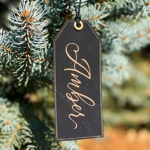 May include: Black leather gift tag with a gold inscription that reads "Huckleberry". The tag is hanging from a blue spruce tree.
