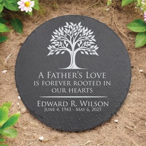May include: A round, black memorial stone with white text and a tree design. The text reads "A Father's Love Is Forever Rooted In Our Hearts" and includes a name and dates. Surrounded by greenery and flowers.