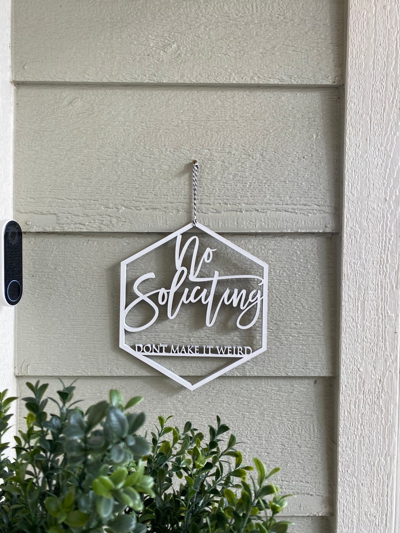 May include: White hexagon-shaped sign with the text "No Soliciting Don't Make It Weird" hanging on a light green wall.