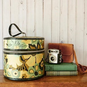 Könnte beinhalten: Eine Vintage-Hutbox mit einem Blumenmuster in Gelb, Grün und Braun, mit einem schwarzen Griff und Reißverschluss. Daneben eine kleine Kamera, eine braune Ledertasche und zwei grüne Bücher.