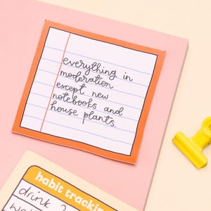May include: A pink and orange notecard with the text "everything in moderation except new notebooks and house plants." on lined paper. A yellow binder clip is on a light yellow surface.