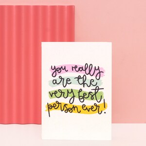 May include: A white greeting card with the handwritten phrase "you really are the very best person ever!" in black ink. The text is layered over horizontal brushstrokes of pink, green, and yellow. A textured coral-coloured panel is on the left.