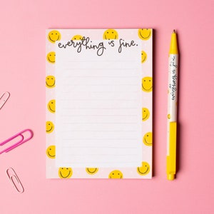 May include: A pink notepad with a white lined interior and a yellow smiley face border. The notepad has the text "everything is fine." written on the top. A yellow pen with the same text is next to the notepad.