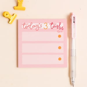 May include: Pink notepad with the text "today's 3 tasks" and three blank lines with orange dots to the right of each line. The notepad has the word "done" written below the title. A white pen and two yellow paper clips are also in the image.