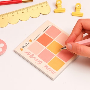 May include: A pink and yellow notepad with a grid pattern and the text "win.  prize.  bring.  run."  A person is writing on the notepad with a pink pen.  There is a gold ruler, two yellow binder clips, and a pink flower button.