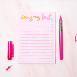 May include: A pink notepad with the text "doing my best" in yellow and pink lettering. The notepad has a lined interior with checkboxes. A pink pen and a pink push pin are also pictured.