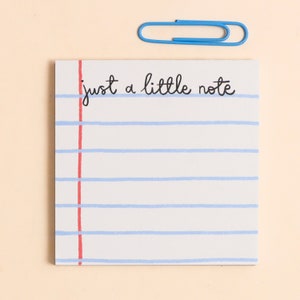 May include: A white notepad with blue horizontal lines and a red vertical line on the left. The words "just a little note" are written in black script. A blue paperclip is positioned above the notepad.