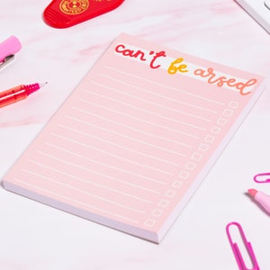 May include: Pink notepad with the text "can't be arsed" in yellow and orange lettering. The notepad has lined paper and a checklist.