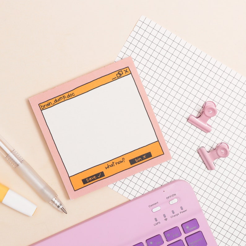 May include: A pink notepad with a computer window design. The notepad has the text "brain_dump.doc" and buttons that say "Save", "what now?" and "bin x". The notepad is on top of a sheet of graph paper with two pink paper clips.