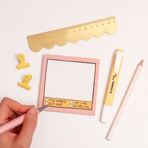 May include: A pink notepad with a dotted pattern and a yellow border. The notepad has a checklist with the options "very important, don't forget", "just a note", and "for another day". There is a yellow ruler with a scalloped edge, a yellow highlighter, and a wooden pencil on a white background.