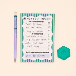 May include: A rectangular daily planner notepad with green and purple vertical stripes, featuring sections for priorities and tasks with handwritten notes, accompanied by a light purple mechanical pencil and a teal hexagonal eraser.