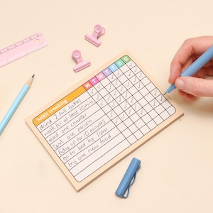 May include: A habit tracking notepad with a grid for daily checkmarks. The notepad has the text "habit tracking" and a list of habits to track. A light blue pen is used to mark the grid. Also, a light blue pencil, a pink ruler, and pink binder clips are on the surface.