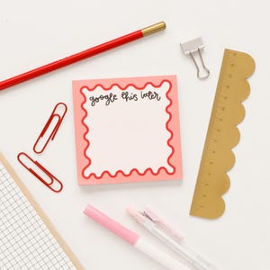 May include: A pink sticky note with a red scalloped border. The note says "google this later". The note is surrounded by office supplies, including a red pencil, gold ruler, and paper clips.