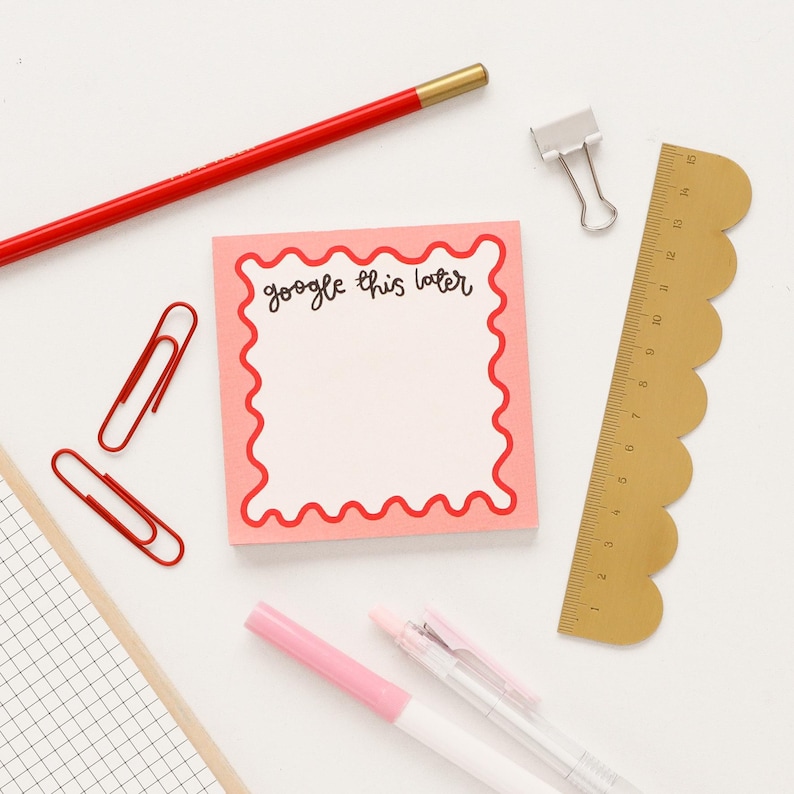 May include: A pink sticky note with a red scalloped border. The note says "google this later". The note is surrounded by office supplies, including a red pencil, gold ruler, and paper clips.