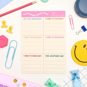 May include: A cream-coloured notepad with pink accents and the text "THE ULTIMATE BRAIN DUMP". It features sections for notes and is surrounded by stationery items, including a pink pencil, paper clips, and a yellow smiley face coaster.