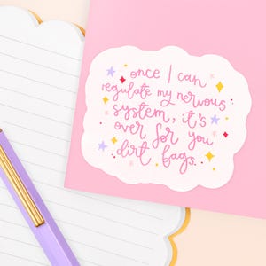 May include: A pink sticker with the text "once I can regulate my nervous system, it's over for you dirt bags." The sticker has a white cloud-like border and is decorated with stars and diamond shapes. A purple pen and lined paper are in the image.
