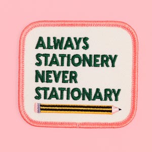 May include: A square embroidered patch with rounded corners featuring a coral pink border and an off-white background. The text reads ALWAYS STATIONERY NEVER STATIONARY in dark green thread, with a yellow and black pencil graphic below.