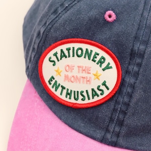 May include: A navy blue baseball cap with a pink brim and a pink eyelet. An oval patch is stitched onto the front featuring a red border, white background, and green text reading "STATIONERY OF THE MONTH ENTHUSIAST" with two yellow stars.