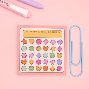 May include: A pink sticky note with the text "for the next 30 days I am going to:" and colourful shapes numbered 1-30. A light blue paper clip and pink and purple pens are also in the image.