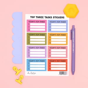 May include: A sheet of Top Three Tasks stickers featuring eight colourful rectangular labels with numbered lines. The sheet includes a light purple scalloped ruler, two yellow binder clips, a yellow hexagon, and a purple pen with text.