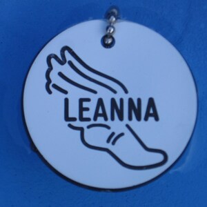 May include: White round tag with a black outline of a running figure and the name "LEANNA" in black text.