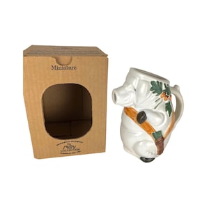 May include: A white ceramic pig-shaped pitcher with a brown handle and spout. The pig has a brown strap across its body and green and orange leaf accents. The pitcher is next to a cardboard box with the word "miniature" printed on it.