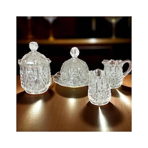 May include: A set of four clear, cut-glass serving dishes. The set includes a lidded jar, a covered butter dish, a small creamer pitcher, and a small open dish. The dishes are arranged on a wooden surface.