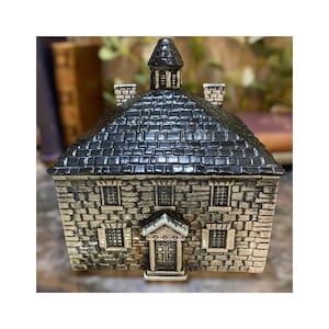 May include: A ceramic house-shaped trinket box with a black tiled roof and a stone-like facade. The miniature building has multiple windows, a front door, and a small cupola on top. The overall design is reminiscent of a historic building.