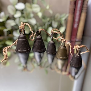 May include: A set of five graduated metal bells in varying shades of dark brown and brass, each featuring etched linear patterns. The bells are connected by a single, thin, light-colored braided cord that loops through the top ring of each bell.