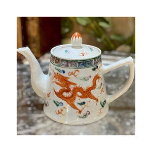 May include: White ceramic teapot with a lid, featuring a vibrant orange dragon design. The teapot has a curved spout and handle, with a decorative border around the top. The lid is topped with a small orange knob. The teapot is decorated with colorful accents.