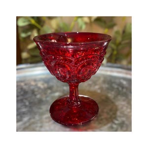 May include: A ruby red, vintage-style glass goblet. The bowl of the glass features a detailed, embossed pattern. The stem and base are also red and have a textured appearance. The glass is likely used for serving beverages.