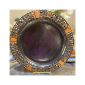 May include: A decorative, round, bronze-colored plate with a dark purple center. The rim features a textured, geometric design with orange-brown accents. The plate is displayed on a gold-colored stand, showcasing its vintage aesthetic.