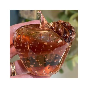 May include: A decorative glass apple with a stem and leaf detail. The apple is a translucent amber color with small air bubbles throughout. The stem and leaf are a darker shade of amber. A unique home decor item.