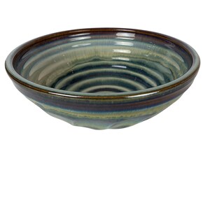 May include: A ceramic bowl with a glossy finish. The bowl features a dark brown rim that transitions to shades of blue, green, and brown. The interior has concentric circles, and the bowl is set against a white background.