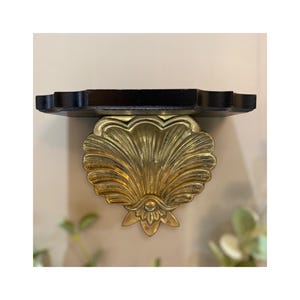 May include: A decorative wall shelf with a black, scalloped top and a gold-colored, seashell-shaped bracket. The bracket has a detailed, textured design. The shelf is mounted on a neutral-colored wall.