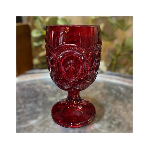 May include: A ruby red glass goblet with a detailed, embossed pattern. The goblet features a round bowl, a short stem, and a circular base. The design includes repeating circular and oval shapes. The glass has a glossy finish and is set against a blurred background.