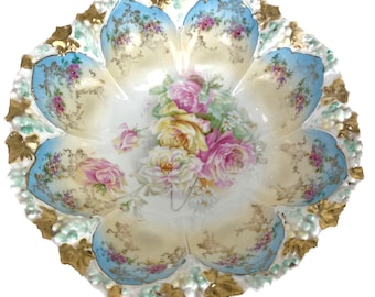 May include: A large, white porcelain bowl with a scalloped edge and a floral design. The bowl is decorated with pink, yellow, and white roses, as well as green leaves. The bowl has a blue and gold rim.