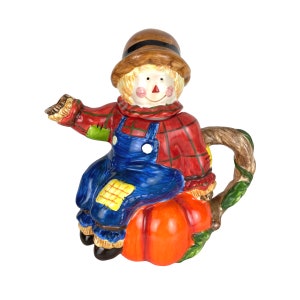 May include: A ceramic teapot in the shape of a smiling scarecrow sitting on an orange pumpkin. The scarecrow wears a brown hat, red plaid shirt, blue overalls, and has a straw-like head. The handle is a brown vine.
