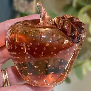 May include: A decorative glass apple in shades of amber and peach. The apple features a stem and a leaf-like design on top. The glass has a bubbly texture, adding visual interest to the piece. A unique home decor item.