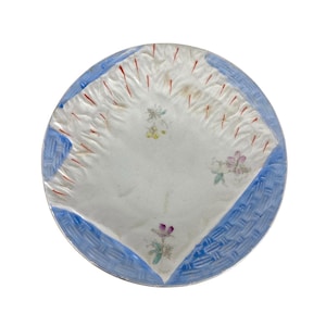 May include: A white ceramic plate with a blue and red rim. The plate has a raised design of a woven pattern and a floral design in the center.