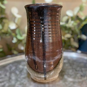 May include: A cylindrical ceramic vase featuring a glossy dark brown and black glaze with vertical drip patterns. The base transitions into a lighter, speckled beige tone. The surface has a smooth, reflective finish with subtle horizontal ridges.