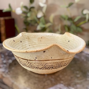 May include: A beige ceramic bowl with a speckled finish and a wavy, scalloped rim. The exterior features a horizontal band of dark brown, hand-drawn wavy line patterns encircling the middle section of the bowl, which has a wide, open shape.