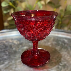 May include: A ruby red glass goblet with a detailed, embossed pattern on the bowl. The goblet has a stem and a circular base. The glass has a vintage aesthetic, and the color is a deep, rich red.